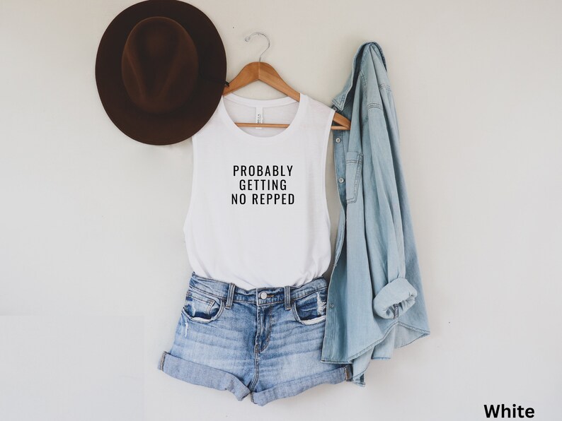 Op de afbeelding: Een wit tanktop zonder mouwen met de tekst "Probably Getting No Repped" erop gedrukt. Het tanktop wordt gemodelleerd door iemand die een spijkerbroek en een spijkerjack draagt.