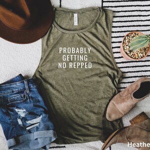 Op de afbeelding: Een groen tanktop zonder mouwen met de tekst "Probably Getting No Repped" in wit. Het tanktop ligt op een gestreept tapijt met een bruine hoed, een spijkerbroekje en een cactus in een pot.