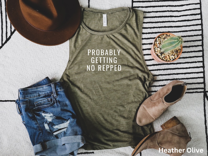 Op de afbeelding: Een groen tanktop zonder mouwen met de tekst "Probably Getting No Repped" in wit. Het tanktop ligt op een gestreept tapijt met een bruine hoed, een spijkerbroekje en een cactus in een pot.