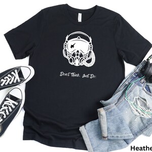 May include: A black t-shirt with a white graphic of a pilot wearing a helmet and oxygen mask. The text "Don't Think, Just Do." is printed below the graphic. The shirt is displayed on a white background with a pair of black sneakers, blue jeans, and headphones.