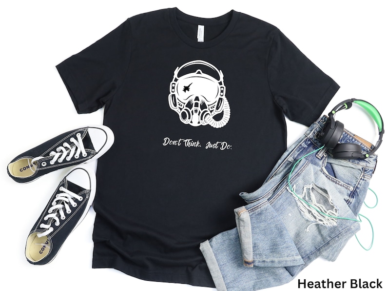 May include: A black t-shirt with a white graphic of a pilot wearing a helmet and oxygen mask. The text "Don't Think, Just Do." is printed below the graphic. The shirt is displayed on a white background with a pair of black sneakers, blue jeans, and headphones.