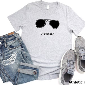 Puede incluir: Una camiseta gris con un gr&aacute;fico negro de gafas de sol y el texto "brewski?" impreso en ella. La camiseta est&aacute; colocada sobre un fondo blanco con un par de pantalones vaqueros azules y zapatillas grises.