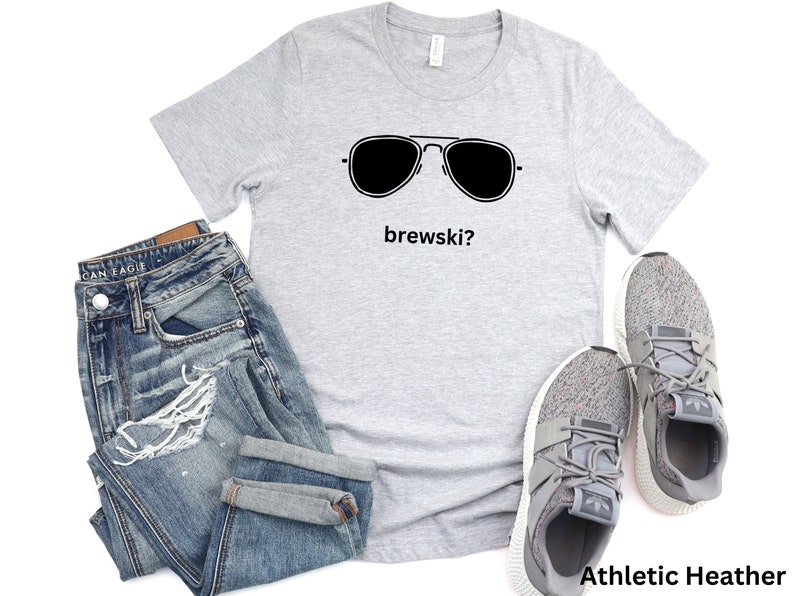 Puede incluir: Una camiseta gris con un gr&aacute;fico negro de gafas de sol y el texto "brewski?" impreso en ella. La camiseta est&aacute; colocada sobre un fondo blanco con un par de pantalones vaqueros azules y zapatillas grises.