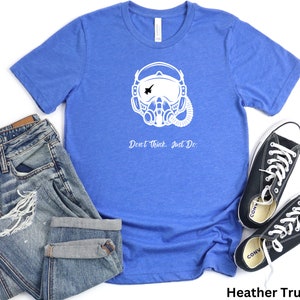 May include: A blue t-shirt with a white graphic of a pilot wearing a helmet and oxygen mask. The text "Don't Think. Just Do." is printed below the graphic. The t-shirt is laid out on a white background with a pair of blue jeans and black sneakers.