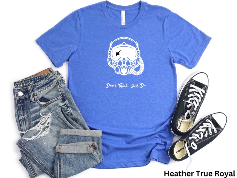 May include: A blue t-shirt with a white graphic of a pilot wearing a helmet and oxygen mask. The text "Don't Think. Just Do." is printed below the graphic. The t-shirt is laid out on a white background with a pair of blue jeans and black sneakers.
