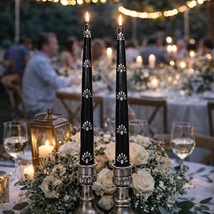 May include: Two lit black taper candles with white floral designs sit in silver candle holders on a table. The candles are surrounded by white roses and greenery. The background features a blurred outdoor setting with string lights and people.