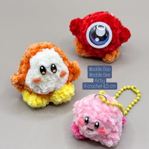 Puede incluir: Tres llaveros de ganchillo de 4,5 cm, incluyendo un Waddle Dee naranja y blanco, un Waddle Doo rojo con un ojo central grande y un Kirby rosa con cadena amarilla. El texto dice: Waddle Doo Waddle Dee Kirby A crochet 4,5 cm.
