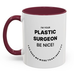May include: White ceramic mug with a claret interior and handle. The mug features the text "I'M YOUR PLASTIC SURGEON BE NICE! YOU NEED ME MORE THAN I NEED YOU" in black, arranged vertically and curved around the mug.
