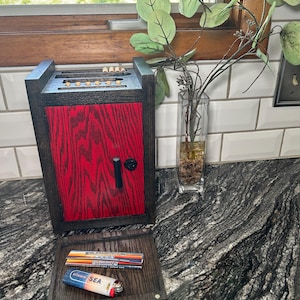 May include: A dark wood storage box with a vibrant red front panel, a black handle, and a circular lock. The top features slots for small items. A matching dark wood tray sits in front, holding a pack of rolling papers and a Vitamin Sea lighter.