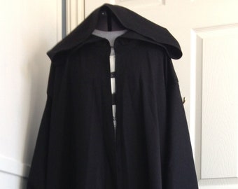 Op de afbeelding: Een zwarte kapmantel met een lange, vloeiende vorm. De mantel is gemaakt van een zachte, gladde stof en heeft een eenvoudige, elegante stijl.
