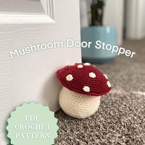 Puede incluir: Un tapón de puerta de ganchillo en forma de seta de color rojo y blanco. La seta está sobre una alfombra marrón junto a una puerta blanca. El texto "PDF CROCHET PATTERN" está sobre una estrella verde.