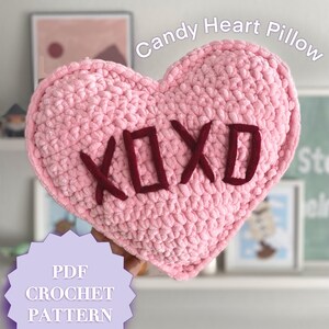 May include: A pink crocheted heart-shaped pillow with the word "XOXO" embroidered in burgundy. The pillow is being held by a hand and the text "PDF CROCHET PATTERN" is visible in the lower left corner of the image.
