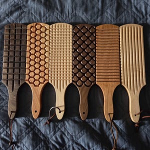 May include: Six wooden gnocchi boards arranged in a row, each featuring a distinct carved pattern including grids, hexagons, scales, and horizontal or vertical lines. Each paddle has a handle with a small hole and a leather hanging cord attached.