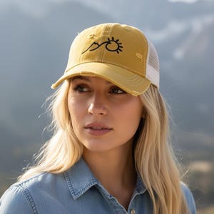 May include: Mustard yellow baseball cap with a distressed finish and a white mesh back. The cap features a black embroidered design of a mountain range and sun. The brim is curved, and the cap is worn by a person.