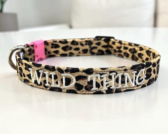 May include: A pet collar featuring a brown and black leopard print pattern with the text WILD THING embroidered in white thread. The collar includes a bright pink plastic buckle and a silver metal D-ring for attachment.