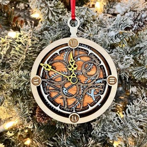 May include: Wooden Christmas ornament in the shape of a clock. The clock face features intricate gear designs in brown, blue, and gold. The ornament hangs from a red ribbon and is displayed on a Christmas tree.