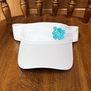 May include: A white visor with a turquoise hibiscus flower embroidered on the left side. The visor is made of a soft material and has a curved brim. The visor is sitting on a wooden chair.