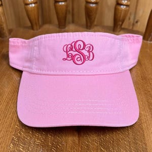 May include: A light pink visor featuring an embroidered monogram in a darker shade of pink on the front panel. The visor has a curved brim with visible stitching and an adjustable strap at the back, constructed from a solid cotton-like fabric.
