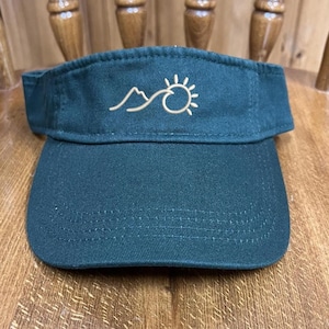 May include: A dark green visor with a curved brim. The visor features a stitched design of a mountain range and a sun in a light tan color. The visor is made of a durable fabric and is designed to shield the eyes from the sun.