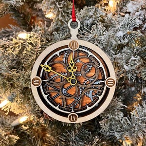May include: A circular wooden ornament featuring a layered clock face design with intricate cut-out gears in silver and brown tones. The piece includes gold-colored ornate clock hands and Roman numerals at the twelve, three, six, and nine positions.