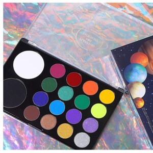 May include: A black eyeshadow palette with a variety of vibrant colors, including pink, green, blue, and yellow. The palette has a clear lid with a planet design. A second palette with planet illustrations is visible.