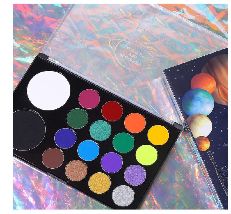 May include: A black eyeshadow palette with a variety of vibrant colors, including pink, green, blue, and yellow. The palette has a clear lid with a planet design. A second palette with planet illustrations is visible.