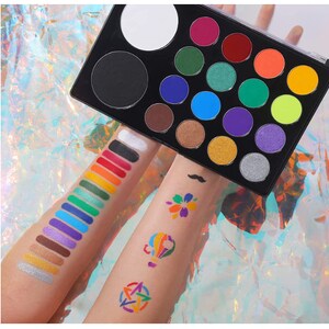 May include: A black makeup palette with a variety of colorful eyeshadows and two large empty pans. Swatches of various colors are displayed on the arms, along with small colorful designs. The background is a shimmering, iridescent surface.