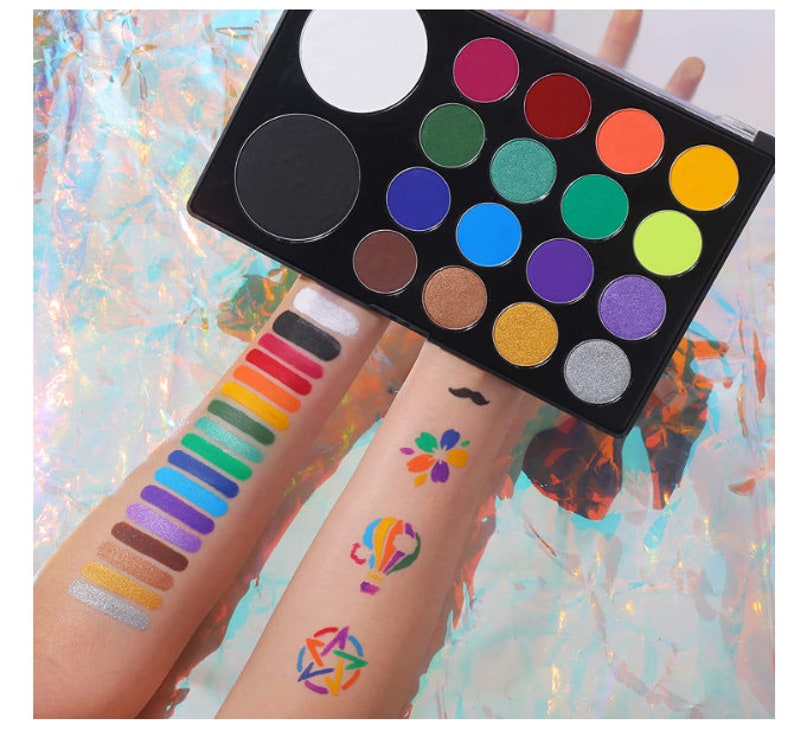 May include: A black makeup palette with a variety of colorful eyeshadows and two large empty pans. Swatches of various colors are displayed on the arms, along with small colorful designs. The background is a shimmering, iridescent surface.