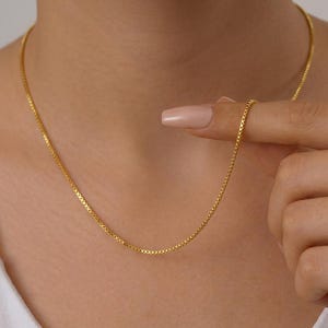 May include: A gold-toned box chain necklace featuring a series of interconnected square links that form a smooth, continuous strand. The necklace rests against the neck, showcasing a polished metallic finish and a uniform, flexible design throughout.