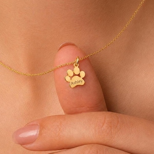 May include: A gold-colored necklace featuring a small, flat pendant shaped like a paw print. The pendant is engraved with the name Ashley in a simple, clear font. The delicate chain connects to the top of the paw print charm, which hangs centered.
