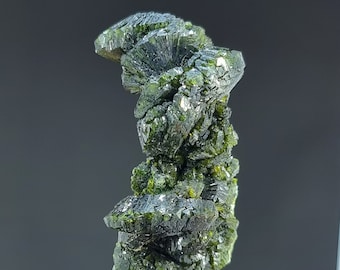May include: A dark green mineral specimen with a layered, crystalline structure. The mineral has a deep, almost black color in some areas, with lighter green highlights. The specimen is mounted on a clear base, showcasing its natural form.