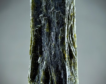 May include: A vertical, elongated mineral specimen with a dark, forest green color and a fibrous, striated texture. The crystal features a flat, rectangular top surface and a rough, uneven base, displaying a complex, layered structure throughout.