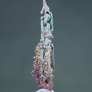 Peut inclure: Gros plan sur un spécimen minéral de forme allongée et unique. Le minéral présente un mélange de couleurs, notamment le sarcelle, le cuivre et le violet. La base est de couleur bleu clair et la texture générale semble rugueuse et cristalline.