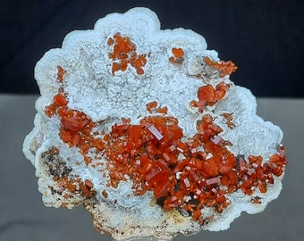 May include: A cluster of bright orange crystals on a white, textured rock. The crystals are arranged in a random pattern and have a shiny, metallic finish.