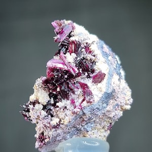 May include: A cluster of deep red, purple, and white crystals on a gray matrix. The crystals are sharp and have a metallic sheen.