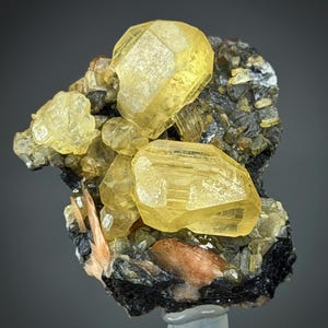 Puede incluir: Un grupo de minerales cristalinos amarillos con aspecto facetado. Los cristales se asientan sobre una matriz de roca gris oscuro y negra, con toques de naranja y marrón. La composición general es un espécimen geológico natural.