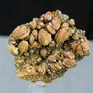 May include: A cluster of golden-brown mineral formations with a crystalline structure. The specimen features rounded, layered formations and a sparkling, textured surface. The mineral is mounted on a clear stand against a dark background.