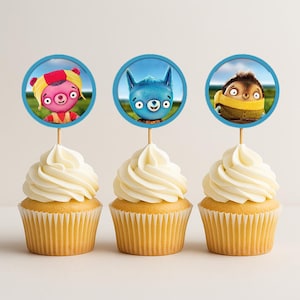 May include: Three cupcakes with white frosting and yellow cake. Each cupcake has a circular topper with a cartoon character. The toppers feature a pink bear, a blue alien, and a brown creature. The toppers are on wooden sticks.