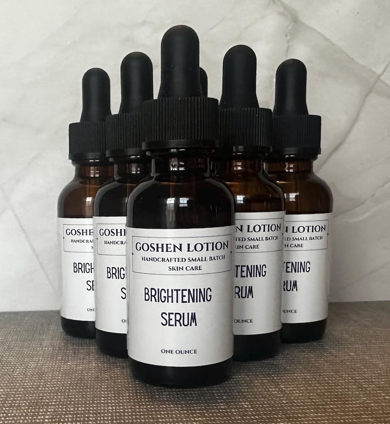 K&ouml;nnte beinhalten: Sechs bernsteinfarbene Glasflaschen mit schwarzen Tropfern, gef&uuml;llt mit einer klaren Fl&uuml;ssigkeit. Die Flaschen sind mit wei&szlig;em Text beschriftet, der "Goshen Lotion", "Handcrafted Small Batch Skin Care", "Brightening Serum", "One Ounce" und "Brightening Serum" lautet.