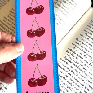 May include: A pink bookmark with a blue border features four illustrations of red cherries with green stems. The text "les cerises" is printed in black at the bottom of the bookmark.