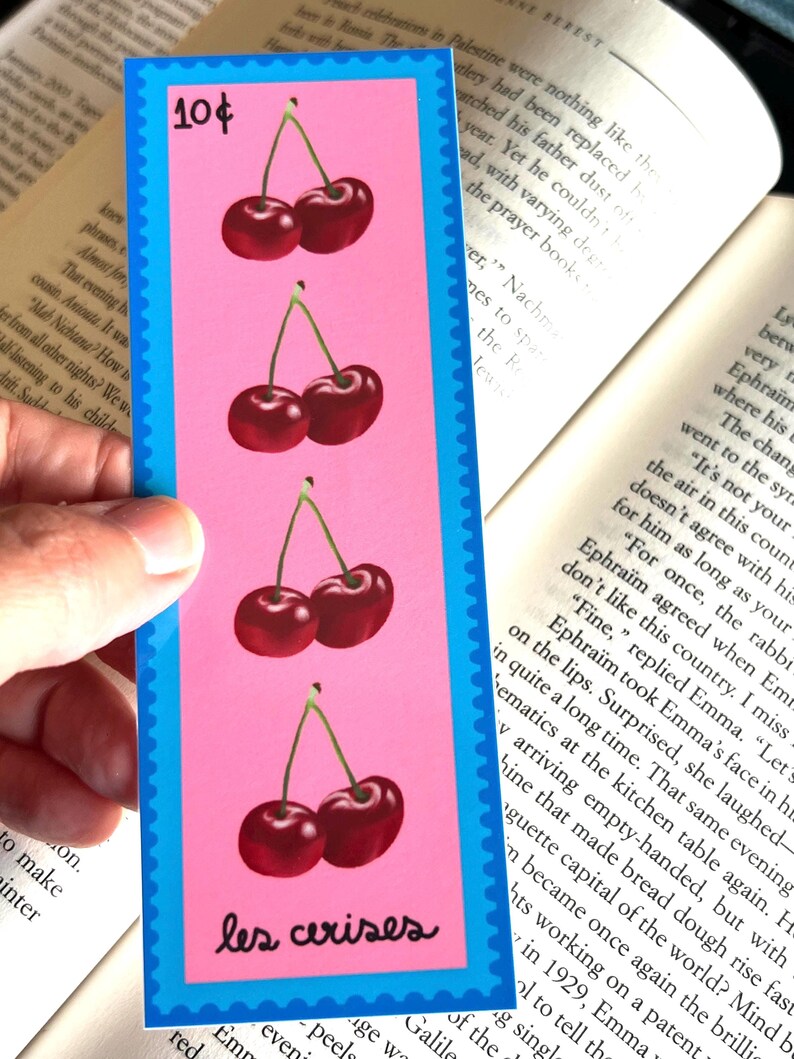 May include: A pink bookmark with a blue border features four illustrations of red cherries with green stems. The text "les cerises" is printed in black at the bottom of the bookmark.