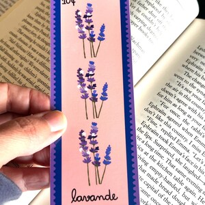 May include: A lavender bookmark with a pink background and a blue border. The bookmark features three sprigs of lavender with purple flowers and white accents. The text "10&cent;" is printed in the top left corner and "lavande" is printed in the bottom right corner.