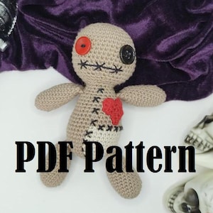 Puede incluir: Patrón de crochet para un muñeco vudú con un ojo de botón rojo, un ojo de botón negro y un corazón rojo en el pecho. El muñeco está tumbado sobre una superficie blanca con dos cráneos blancos en el fondo. El texto "PDF Pattern" está impreso en negro sobre la superficie blanca.