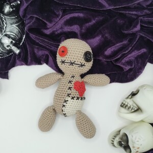 Puede incluir: Una mu&ntilde;eca vud&uacute; de crochet con un ojo de bot&oacute;n rojo, un ojo de bot&oacute;n negro y un coraz&oacute;n rojo cosido en el pecho. La mu&ntilde;eca est&aacute; tumbada sobre una superficie blanca con una tela de terciopelo morado en el fondo.