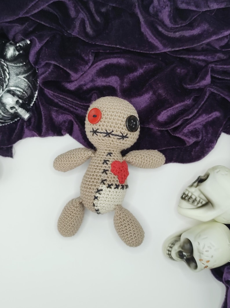 Puede incluir: Una mu&ntilde;eca vud&uacute; de crochet con un ojo de bot&oacute;n rojo, un ojo de bot&oacute;n negro y un coraz&oacute;n rojo cosido en el pecho. La mu&ntilde;eca est&aacute; tumbada sobre una superficie blanca con una tela de terciopelo morado en el fondo.
