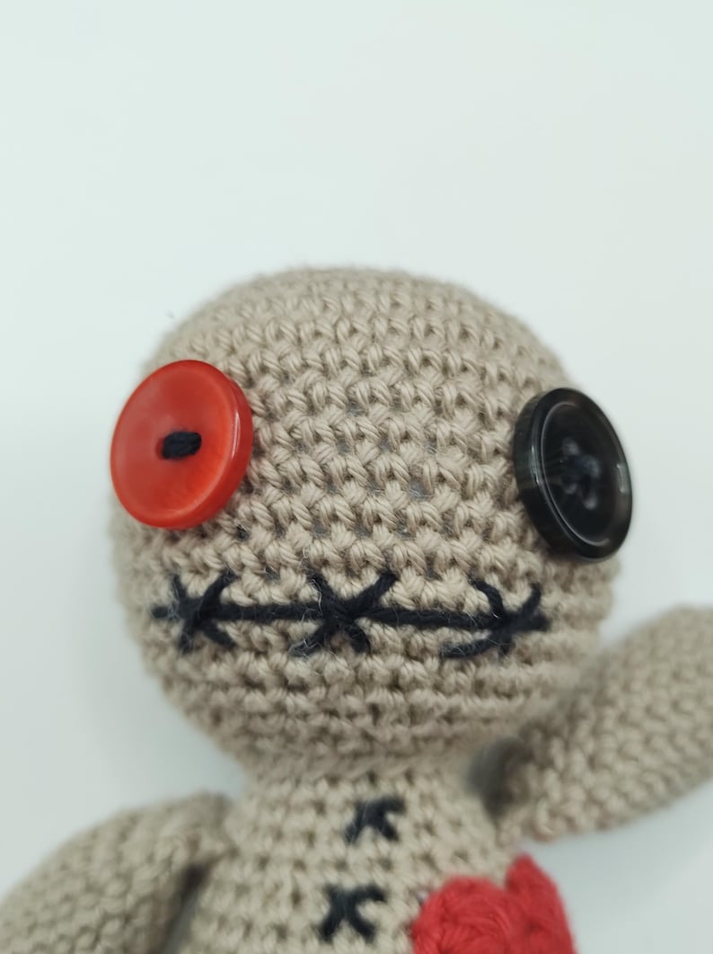 Puede incluir: Una mu&ntilde;eca vud&uacute; de crochet con un bot&oacute;n rojo para un ojo, un bot&oacute;n negro para el otro ojo y una boca cosida negra. La mu&ntilde;eca lleva un coraz&oacute;n rojo.