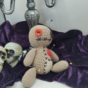 Puede incluir: Una mu&ntilde;eca vud&uacute; de crochet con un ojo de bot&oacute;n rojo, un coraz&oacute;n rojo y varios alfileres que sobresalen. La mu&ntilde;eca est&aacute; sentada sobre una tela de terciopelo morado con un candelabro plateado en el fondo.