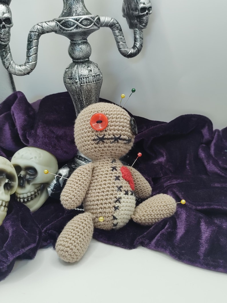Puede incluir: Una mu&ntilde;eca vud&uacute; de crochet con un ojo de bot&oacute;n rojo, un coraz&oacute;n rojo y varios alfileres que sobresalen. La mu&ntilde;eca est&aacute; sentada sobre una tela de terciopelo morado con un candelabro plateado en el fondo.