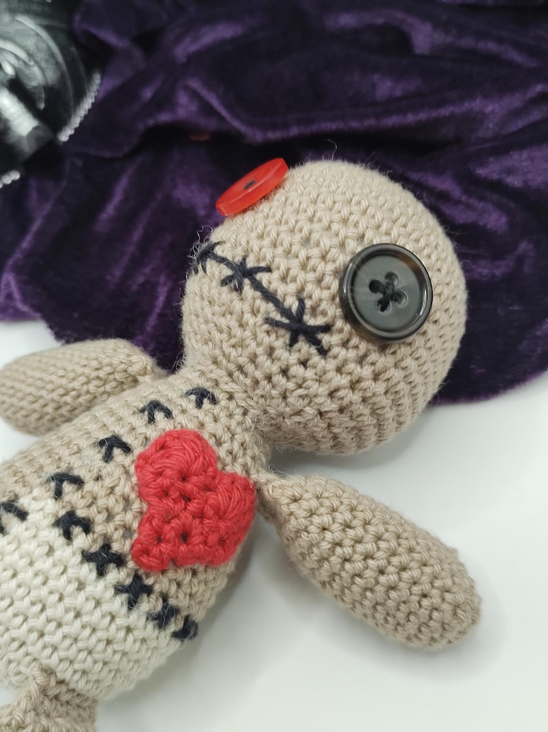 Puede incluir: Una mu&ntilde;eca vud&uacute; de crochet con un coraz&oacute;n rojo en el pecho, ojos de bot&oacute;n negro y nariz de bot&oacute;n rojo.