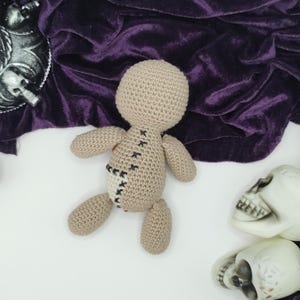 Puede incluir: Una mu&ntilde;eca vud&uacute; de crochet con costuras negras y un parche de tela marr&oacute;n en la espalda. La mu&ntilde;eca est&aacute; tumbada sobre una superficie blanca con dos cr&aacute;neos de pl&aacute;stico blanco.